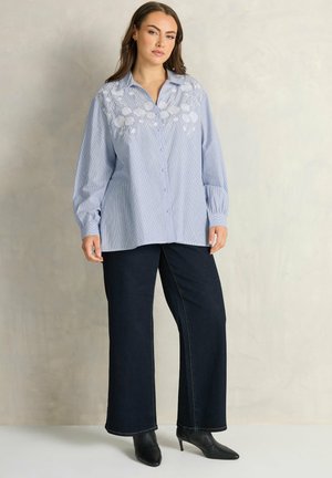 Kvinde iført en lyseblå skjorte med hvide blomsterbroderier og mørke, brede jeans, stående foran en ensfarvet beige baggrund.