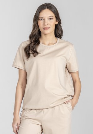 Kobieta o długich brązowych włosach, ubraną w beżową koszulkę z krótkim rękawem i pasujące spodnie, stojąca z jedną ręką w kieszeni na szarym tle.