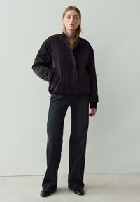Jeune femme portant une veste noire rembourrée, un jean large noir et des chaussures noires, se tenant devant un fond gris clair uni.