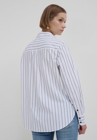 Chemise blanche à manches longues avec des fines rayures noires, pli au dos, ourlet courbé et poignet boutonné, portée avec un jean bleu.