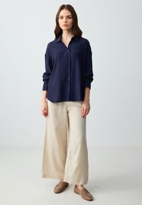 Marineblauw overhemd met lange mouwen, crèmekleurige broek met wijde pijpen en bruine instapschoenen, tegen een lichte achtergrond.
