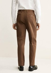 Pantaloni marroni con un taglio su misura, dotati di due tasche posteriori, una texture liscia e una piega frontale, abbinati a scarpe nere.