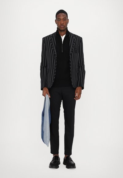 Black and white pinstripe blazer, black zip-up sweater, black trousers, and black shoes; holding a light blue denim jacket.