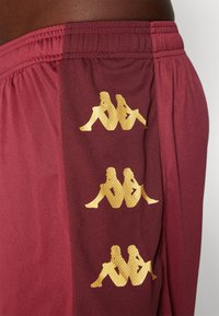 Pantalones cortos deportivos granate hechos de tela sintética, con tres logotipos bordados en dorado en el costado, textura suave y cinturilla elástica.