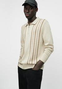 Beige Strickpullover mit einem Reißverschlusskragen, der vertikale braune Streifen aufweist. Aus weichem Stoff gefertigt und kombiniert mit schwarzen Jeans.