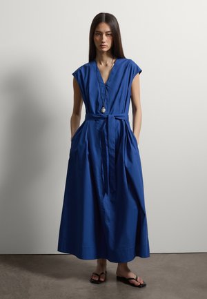 Donna con abito lungo blu senza maniche con cintura annodata in vita e sandali neri, in piedi davanti a una parete chiara e uniforme.