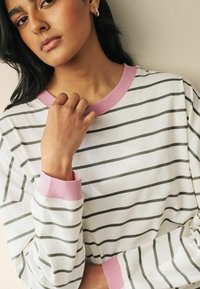 Vit sweatshirt med gröna ränder, rosa ribbat krage och ärmslut, med en avslappnad passform och mjuk textur. Armen är höjd nära nacken.
