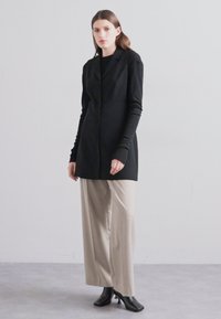 Melna pielāgota garā žakete ar vienu pogu, saskaņota ar bēšiem plašiem biksēm un melniem potīšu zābakiem. Gluda auduma un minimāla dizaina.