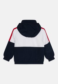 Navyblau-weiße Kapuzenjacke mit roten Akzenten, ausgestattet mit elastischen Bündchen und einem gerafften Saum. Hergestellt aus einem leichten, glatten Stoff.