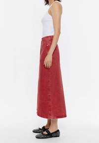 Rode denim maxi-rok met een hoge taille, zijzakken en een rechte snit; gecombineerd met zwarte platte schoenen met studs en een wit hemdje.