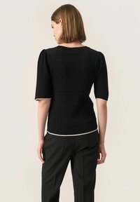 Černý pletený top s texturovaným rib designem, krátkými nabíranými rukávy a kontrastním krémovým lemem kolem výstřihu a spodního okraje.