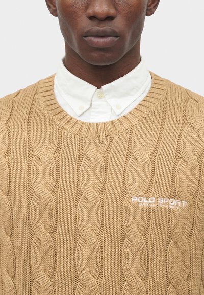 Άνδρας που φορά ένα μπεζ πουλόβερ με πλεξούδα πάνω από ένα λευκό πουκάμισο με κουμπιά και κεντημένο λογότυπο "Polo Sport" στο στήθος.