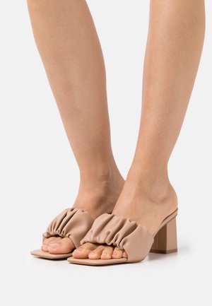 Voeten in beige geplooide leren open teen blokhakken tegen een witte achtergrond.