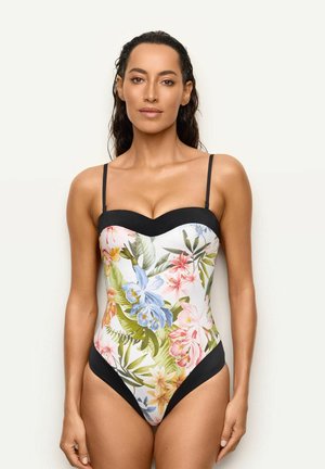 Mujer de pie sobre fondo blanco con un traje de baño de una pieza con flores, con tirantes y ribete negro.