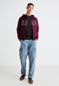 Sudadera con cremallera de color burdeos con el logo de "GAP", camiseta negra debajo, pantalones cargo de color azul claro y zapatos marrones con suelas blancas. Fondo liso.