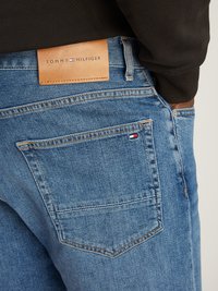 Los vaqueros de mezclilla azules cuentan con una etiqueta de cuero marrón marcada "TOMMY HILFIGER" en la cinturilla, con un pequeño logotipo en el bolsillo trasero.