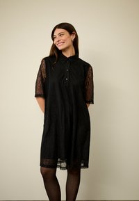 Melna mežģīņu krekla kleita ar apkaklīti, īsām caurspīdīgām piedurknēm un pogu aizdari. Mīksta tekstūra ar plūstošu siluetu un apakšmalas akcentu.