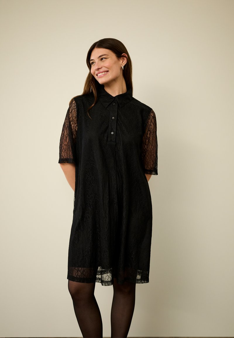 Melna mežģīņu krekla kleita ar apkaklīti, īsām caurspīdīgām piedurknēm un pogu aizdari. Mīksta tekstūra ar plūstošu siluetu un apakšmalas akcentu.