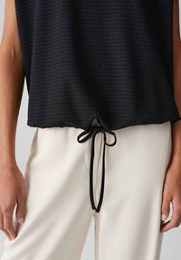 Zwarte textuur top met een trekkoord in de taille, gecombineerd met lichtgekleurde, gladde stofbroeken, waarbij de handen bij de zoom zichtbaar zijn.