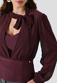 Blusa bordeaux con chiusura a fiocco e maniche trasparenti. Presenta un corpetto aderente con una texture liscia e una leggera lucentezza. Bottone decorativo in vita.