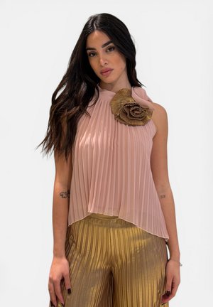 Donna con capelli lunghi e scuri che indossa un top rosa senza maniche plissettato con un fiore di stoffa e pantaloni plissettati metallici dorati.