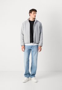 Grå polstret jakke med høy krage, svart skjorte under, lyseblå jeans og hvite joggesko. Glatt stoff, glidelås lukking.