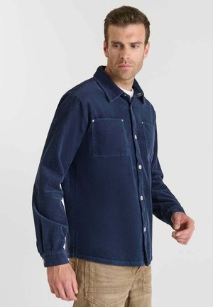 Camisa de pana azul marino con cuello, dos bolsillos en el pecho y cierre frontal de botones, que presenta una textura de acanalado vertical y mangas largas.