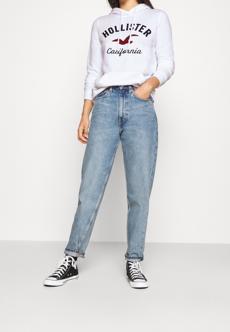 Ljusgrå hoodie med tryck av "Hollister California", kombinerad med högmidjade blå denimjeans och svarta högstövlar.