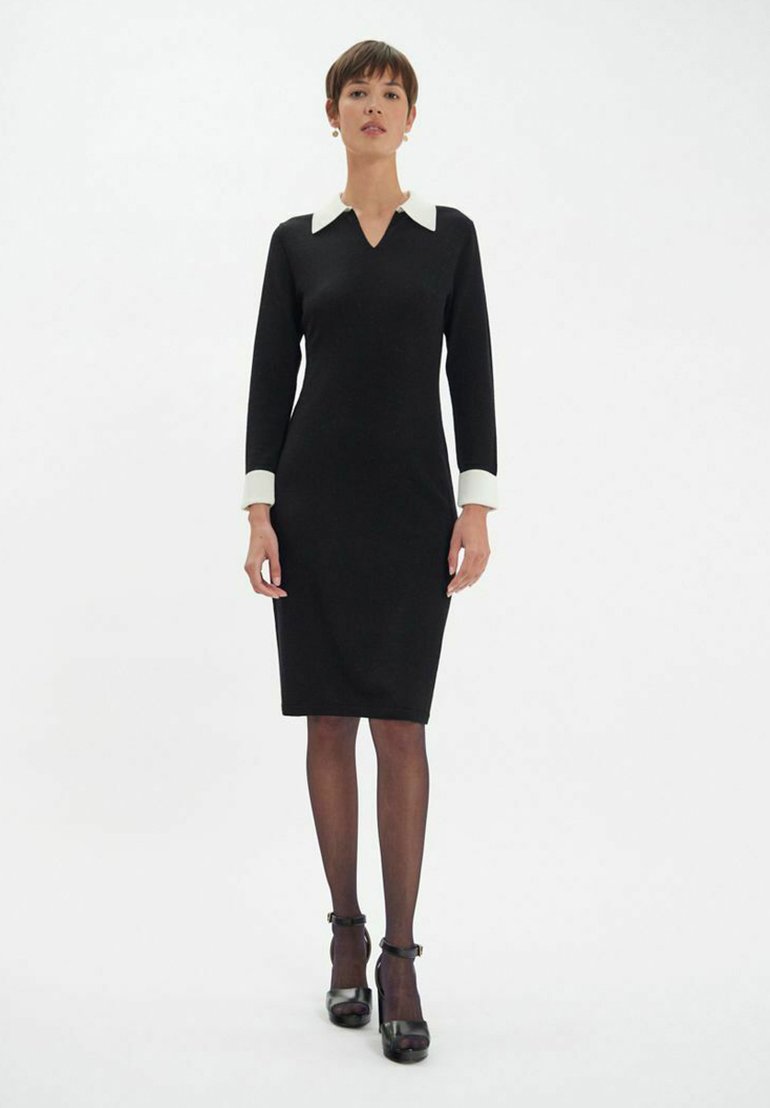 Robe noire ajustée avec un décolleté en V, col blanc et poignets blancs. Le mannequin porte des collants transparents et des escarpins en cuir, se tenant sur un fond uni.