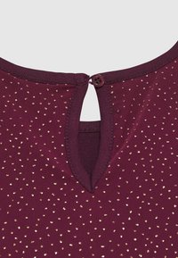 Close-up van bordeauxstof met kleine gouden stippen, met een ronde halslijn met een sleutelgatopening die wordt vastgemaakt met een kleine knop.