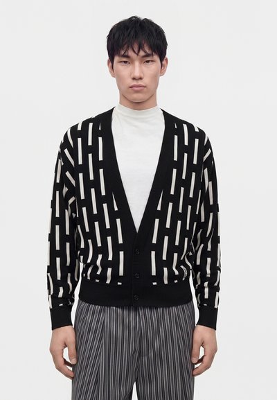 Homme portant un cardigan noir avec des barres blanches verticales sur une chemise blanche et un pantalon gris à rayures verticales, debout devant un fond uni.