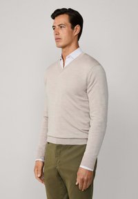 Mężczyzna w beżowym swetrze w serek, noszący białą koszulę z kołnierzykiem oraz oliwkowe spodnie, patrzący lekko na bok na jednolitym tle.