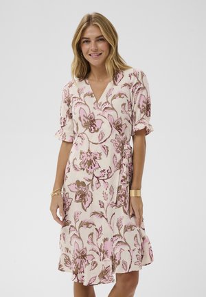 Blondínka sa usmieva, má na sebe ružové a hnedé kvetované zavinovacie šaty s volánmi na rukávoch a spodnom okraji, doplnené zlatými náramkami.