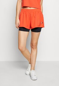 Pantalones cortos deportivos naranjas con un forro interior negro, cintura elástica y un pequeño parche de logo en el costado. Combinados con zapatillas blancas.
