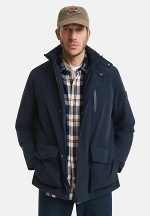 Man som bär en khakifärgad keps, en marinblå jacka över en vit, marinblå och röd rutig skjorta, står med händerna i jackfickorna mot en vit bakgrund.