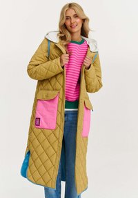Prešívaný dlhý kabát v khaki s ružovými vreckami, šedou kapucňou a modrými akcentmi. Má zapínanie na gombíky a pod ním je pruhovaný sveter s ružovými pruhmi.