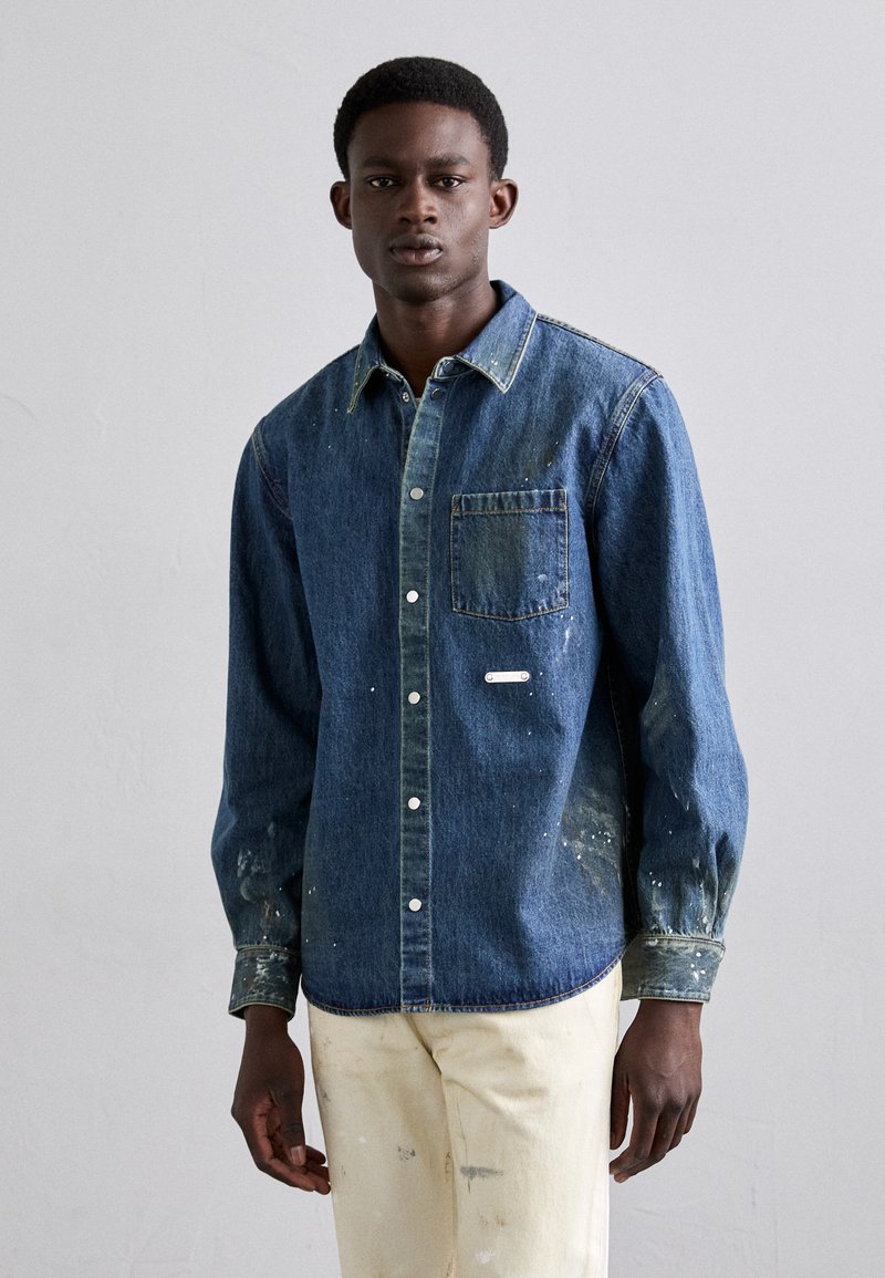 Camisa de mezclilla en azul oscuro con cuello abotonado, bolsillo frontal y manchas de pintura blanca. Mangas largas con puños abotonados.