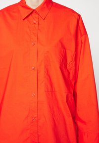 Chemise orange vif à boutons, avec col et poche poitrine, portée par une personne sur un fond uni.