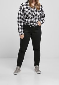Persona che indossa una camicia a quadri bianca e nera infilata in pantaloni neri e sneakers alte grigie su uno sfondo semplice.