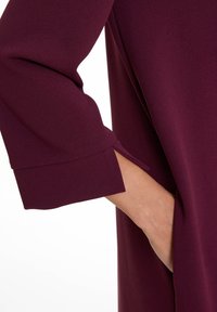 Robe bordeaux en tissu texturé, avec des manches trois-quarts et des poches latérales mises en valeur par des fentes à l'ourlet.