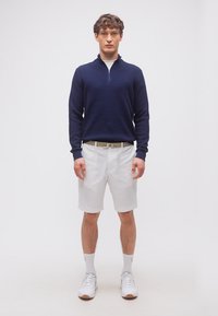 Άνδρας στέκεται φορώντας μπλε σκούρο πουλόβερ με φερμουάρ έως τη μέση, λευκά σορτς, λευκή ζώνη, λευκές κάλτσες και λευκά αθλητικά παπούτσια σε απλό ανοιχτό φόντο.