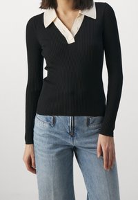 Haut noir à manches longues côtelé avec un col contrastant crème, doté d'une patte de boutonnage à moitié ouverte et d'une coupe ajustée, associé à un jean bleu clair.