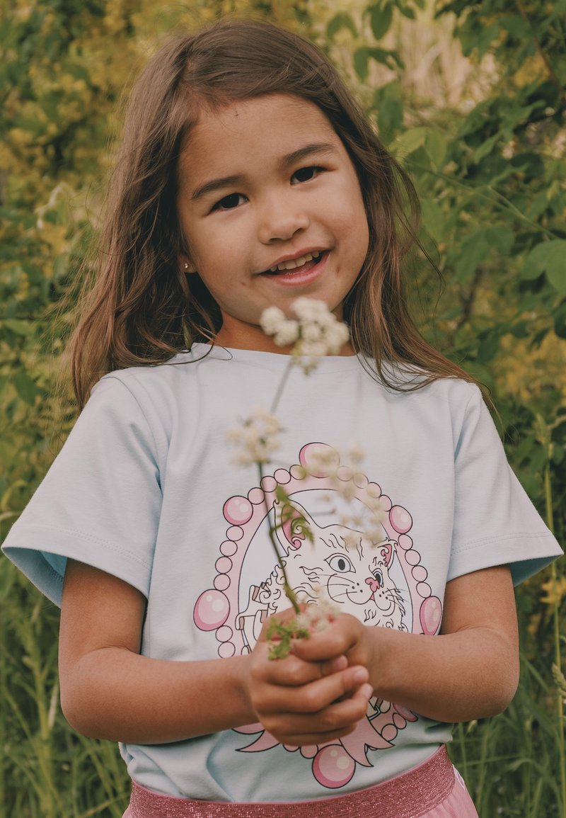 Fată tânără cu păr lung zâmbind, ținând flori sălbatice albe, purtând o cămașă albastru deschis cu o ilustrație de pisică, stând în aer liber în mijlocul naturii verzi.