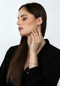 Silberarmband mit herzförmigen und ovalen Anhängern, an einer Hand getragen, vor einem schlichten Hintergrund. Das Modell hat lange Haare und ein schwarzes Shirt.