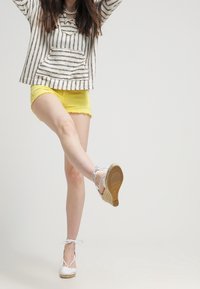 Persona que lleva pantalones cortos amarillos y un top a rayas, levantando una pierna hacia adelante mientras está de pie sobre unas alpargatas de cuña blancas.