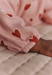 Rosa tyg med röda hjärtmönster och vita vågiga detaljer vid muddarna. En hand vilar på en quiltad beige yta.