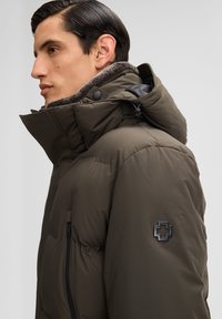 Pufferjack in olijfgroen met een hoge kraag, zachte voering, zijzak met rits en een zwart logo op de schouder.