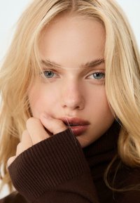 Nahaufnahme eines Gesichts mit blonden Haaren und blauen Augen. Das Motiv trägt einen dunkelbraunen, gerippten Pullover, die Hand ruht am Kinn. Die Hauttextur ist glatt.