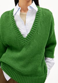 Pull en tricot vert avec un col en V, des manches larges et un ourlet côtelé, superposé à une chemise à col bleu clair, avec un sous-vêtement neutre.
