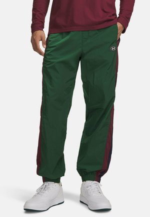 Mann trägt grüne Trainingshose mit burgunderfarbenen Seitenstreifen und weißen Sneakers, eine Hand in der Tasche und ein burgunderfarbenes Langarmshirt.
